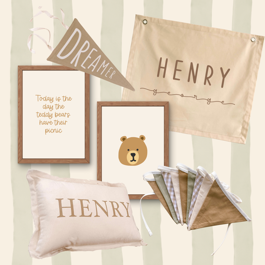 A coordinated nursery decor set featuring a beige fabric banner, a framed quote print, a framed bear illustration print, a personalized cream pillow with the name HENRY, and a folded set of matching fabric flags, all arranged against a soft cream-and-beige striped background.