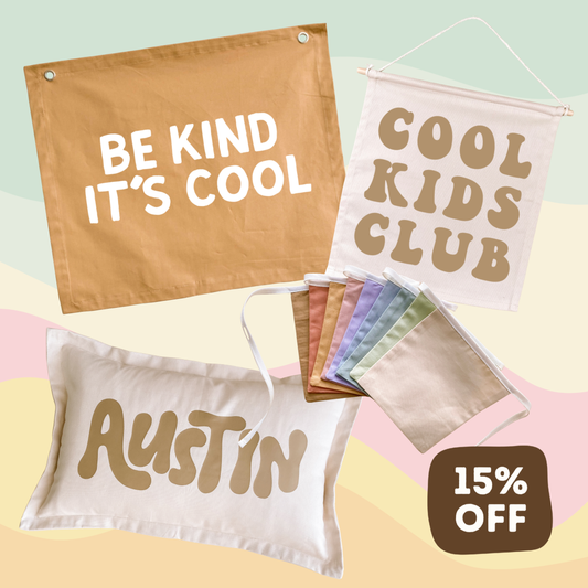 A set of customizable fabric wall flags and matching face masks displayed on a pastel background. The flags include designs reading 'BE KIND IT'S COOL', 'COOL KIDS CLUB', and 'AUSTIN' in brown lettering on white and tan backgrounds.