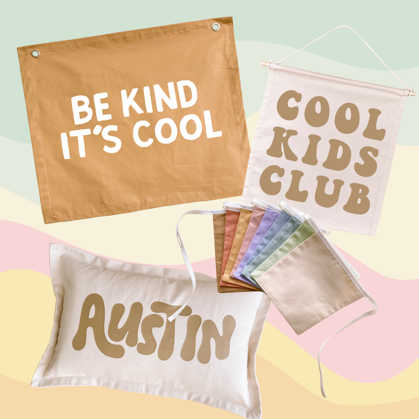 A coordinated set of printed fabric items on a pastel abstract background: a tan banner reading “BE KIND IT’S COOL,” a white hanging banner reading “COOL KIDS CLUB,” a white pillow with “AUSTIN” in tan, and a small stack of folded fabric pieces in assorted pastel and tan colors.