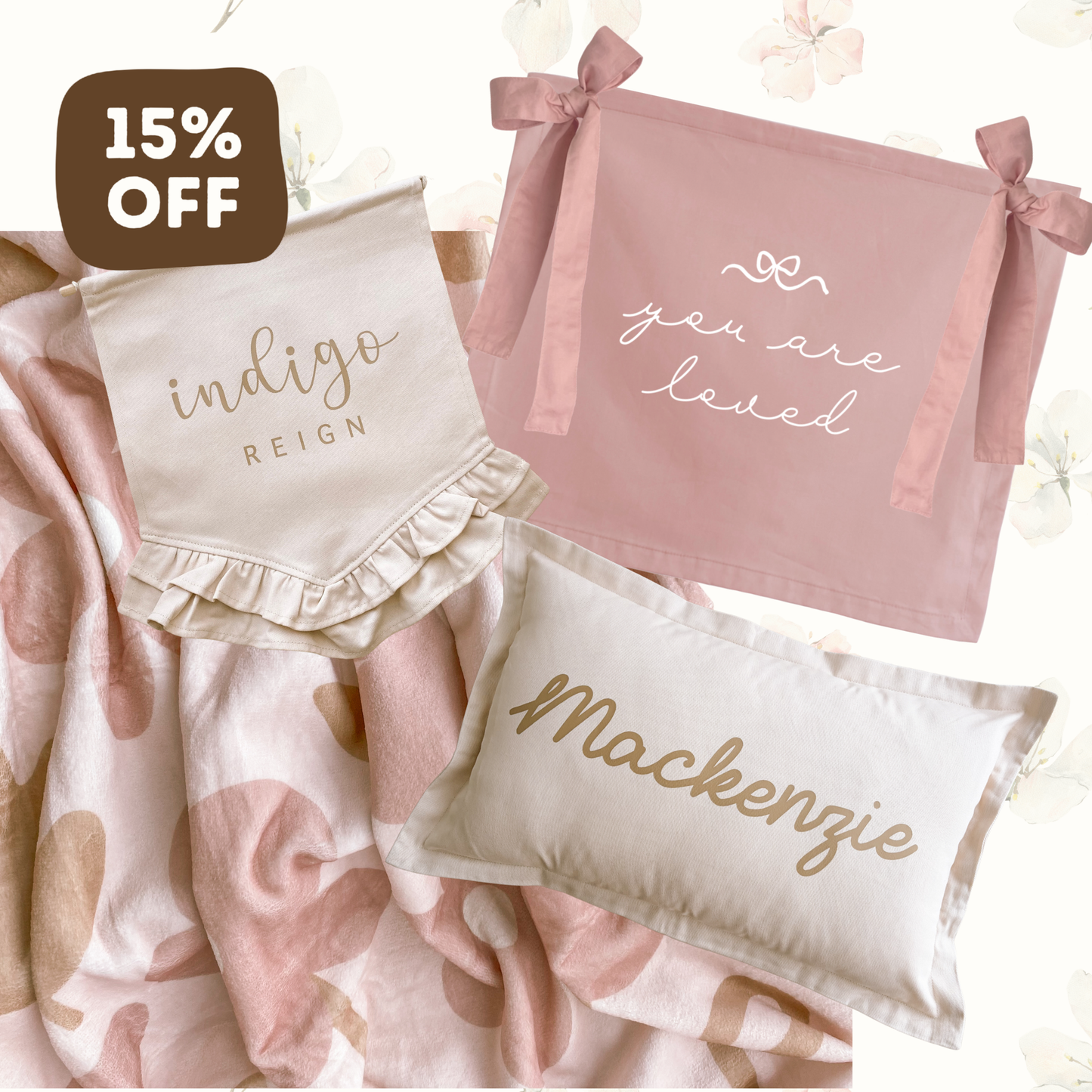 A styled bedding set featuring a pink and white patterned blanket, a pink pillow with 'you are loved' in white script, a cream pillow with 'Mackenzie' in gold script, and a cream bib with 'indigo REIGN' in gold script. A brown '15% OFF' sticker is in the top left corner.