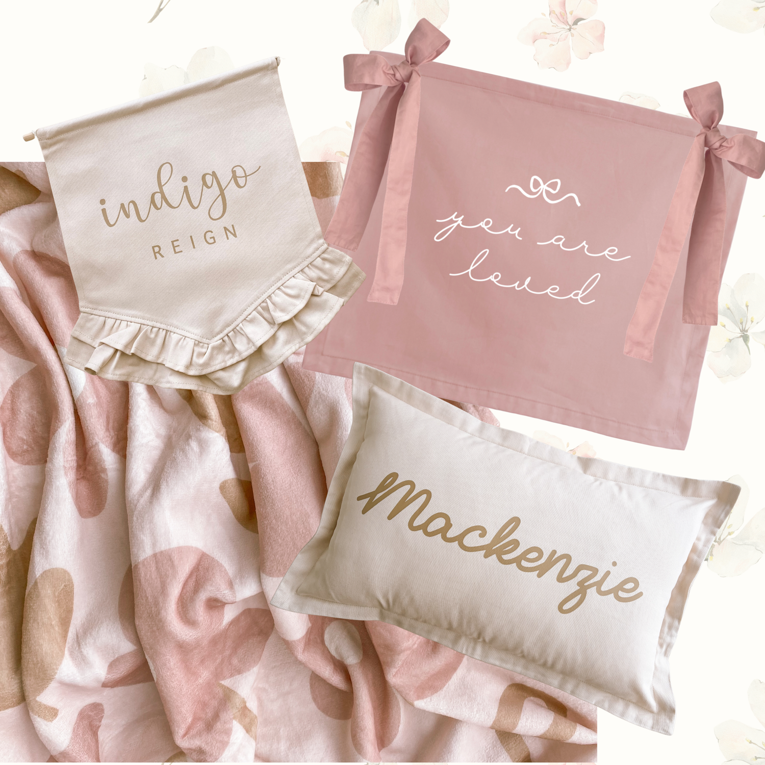 A styled flat-lay of personalized baby bedding and accessories, including a pink floral blanket, a pink bib with 'you are loved' text, a cream pillow with 'Mackenzie' in gold script, and a cream banner with 'indigo REIGN' text.