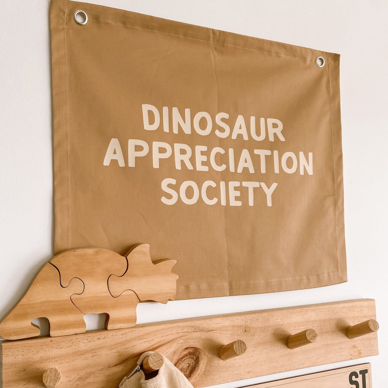 A tan fabric banner reading 'DINOSAUR APPRECIATION SOCIETY' hangs on a white wall above a wooden coat rack with dinosaur-shaped pegs and a beige hat hanging from one peg. A small sign reads 'BEAU ST'.