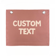 A rectangular dusty rose canvas banner with two silver metal grommets at the top corners and the words 'CUSTOM TEXT' printed in a light beige hand-drawn font, shown on a plain white background.